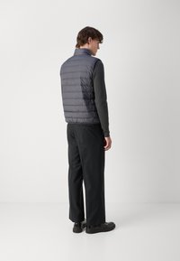Graue Steppweste mit horizontalen Nähten, hohem Kragen und glatter Textur, kombiniert mit schwarzen Hosen und eleganten schwarzen Schuhen.