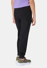 Bambino che indossa pantaloni jogger neri, maglietta viola, calzini bianchi e sneaker verdi con suole bianche, in piedi di lato su uno sfondo bianco.