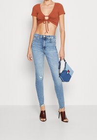 Top corto de punto en color óxido con detalle de nudo, jeans ajustados de tela vaquera claros con rodilla desgastada, zuecos de tacón abierto en color marrón, y un pequeño bolso de mezclilla.