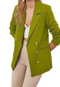 Zielony blezer z gładkiego materiału, dwurzędowy fason z srebrnymi guzikami, z wyciętymi klapami i podwiniętymi rękawami. Nosi się go na biały top.