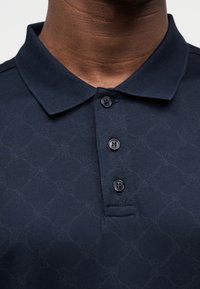 Μπλε σκούρο πόλο μπλουζάκι με διακριτικό γεωμετρικό μοτίβο, διαθέτει γιακά και τρία μαύρα κουμπιά στον λαιμό, κατασκευασμένο από υφασμάτινη ύφανση με υφή.