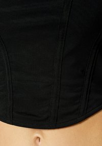 Schwarzes, tailliertes Top aus strukturiertem Stoff, das Nähte und einen geschwungenen Saum aufweist. Das Material wirkt atmungsaktiv und figurbetont.