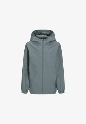 Zöld vízálló dzseki kapucnival, szürke mintás inggel viselve, széles szárú kék farmerrel és barna cipővel párosítva.