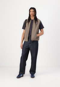 Beige sleeveless vest with a zipper and pockets, worn over a dark t-shirt; paired with loose dark jeans and blue athletic shoes.