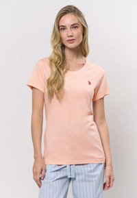 Vaalean persikanvärinen lyhythihainen t-paita, valmistettu pehmeästä kankaasta, jossa pyöreä pääntie ja pieni punainen logo vasemmassa rinnassa.