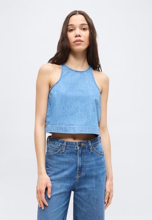 Młoda kobieta o długich ciemnych włosach, ubrana w bezrękawnik z dżinsu z odsłoniętym brzuchem i wysokim stanem niebieskich dżinsów, na jednolitym tle.