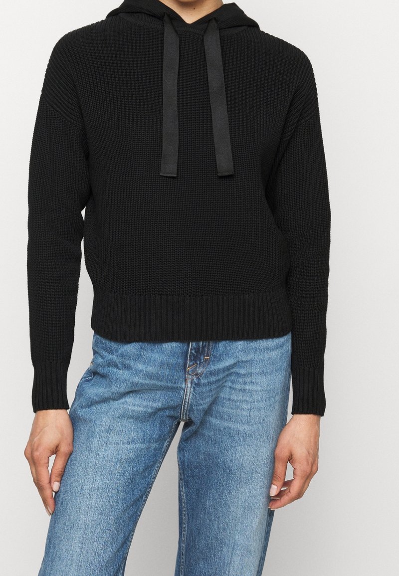 Personne portant un sweat à capuche tricoté noir avec cordons et un jean denim bleu clair, debout devant un fond blanc uni.