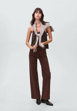 Pantaloni larghi marroni abbinati a un top senza maniche scuro e a una sciarpa grigia chiara drappeggiata sulle spalle. Gli accessori includono collane sovrapposte.