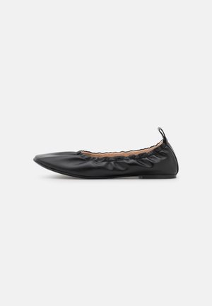 Ballerine plate en cuir noir avec bout pointu, bord élastique froncé et languette à l'arrière, présentée sur un fond uni.