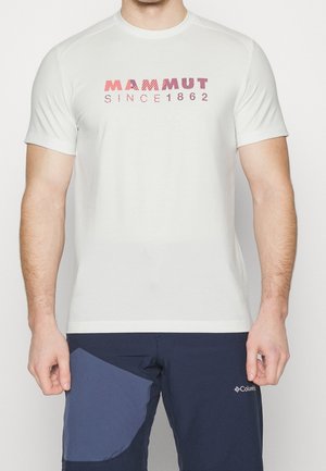 Mężczyzna w białej koszulce Mammut z napisem "SINCE 1862" oraz granatowych spodniach Columbia z jasnoniebieską łatą na lewej udzie.