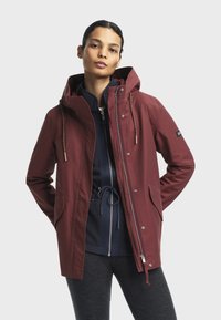 Veste imperméable bordeaux avec capuche, dotée de cordons de serrage, de poches latérales et de zips. Portée par-dessus une veste à doublure bleu foncé.