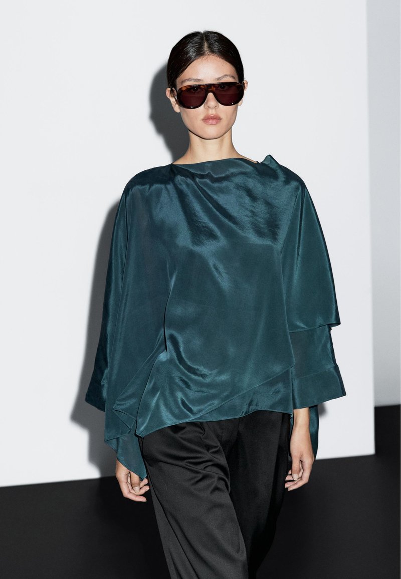 Teal satin-outfit med draperad design och breda ärmar, kombinerad med svarta byxor. Utrustad med stora mörka solglasögon.