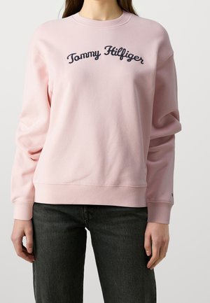 Female wearing light pink sweatshirt with "Tommy Hilfiger" text in navy blue and black jeans, standing against a plain background.