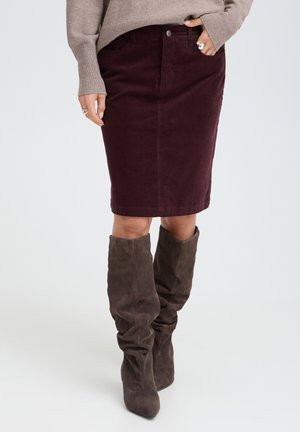 Kvinde iført en mørk burgunderrød nederdel, der går til knæene, taupe sweater og løstsiddende mørkebrune ruskind knæhøje støvler på en ensfarvet lys baggrund.