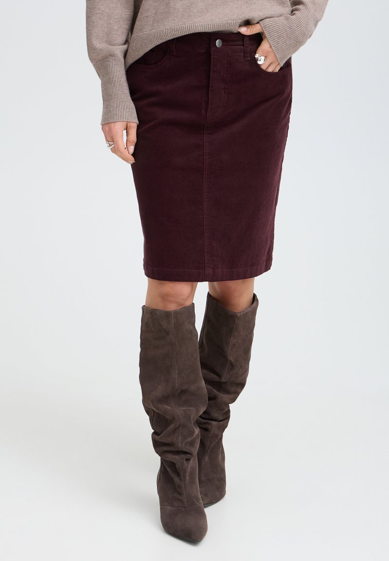 Femme portant une jupe bordeaux foncée longueur genoux, un pull taupe et des bottes molles en daim marron foncé montant jusqu'au genou sur un fond clair uni.