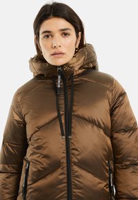 Chaqueta acolchada marrón con capucha, acolchado en forma de chevron, bolsillos con cremallera y cordones negros. El material tiene una textura lisa y brillante.