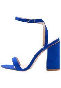 Sininen samettinen korkokenkä, jossa on leveä varvasremmi, nilkkaremmi soljella ja paksu blokiakseli vakauden vuoksi.