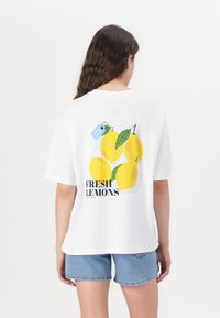 Camiseta de algodón blanca con un gráfico de limones amarillos, una etiqueta azul que dice "100% Natural" y texto negro "LIMONES FRESCOS" en la parte posterior.