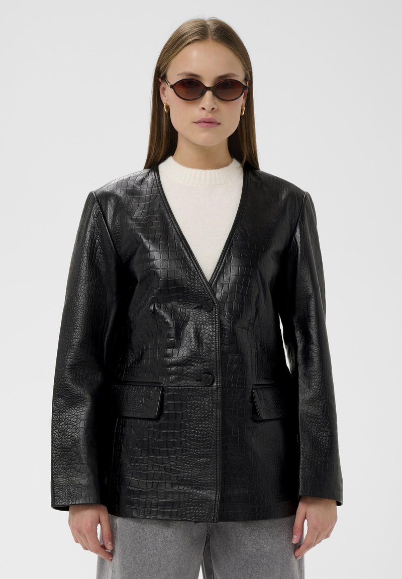 Veste en similicuir noir à texture crocodile, avec un col en V et deux poches avant. Portée sur un pull léger.