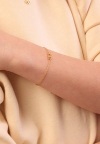 Gouden armband met een hartvormige bedel, delicate kettingontwerp, gedragen om een naakte pols tegen een zachte, beige stoffen achtergrond.