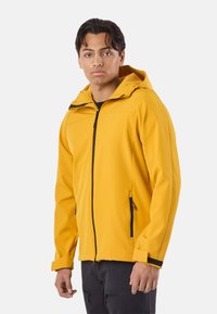 Nuori mies, joka pukeutunut keltaiseen hupulliseen takkiin, jossa on musta vetoketju, ja tummiin housuihin, seisoo yksinkertaisen vaalean taustan edessä ja katsoo eteenpäin.