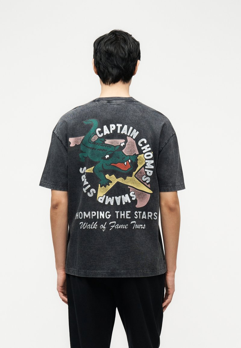 Άτομο που φοράει μια ξεθωριασμένη μαύρη μπλούζα με ένα πολύχρωμο καρτούν αλιγάτορα και κείμενο «Captain Chomps Swamp Stars Romping the Stars Walk of Fame Tours» στο πίσω μέρος.