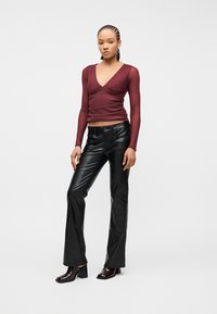 Γυναίκα που στέκεται φορώντας βαθύ κόκκινο μπλουζάκι με μακριά μανίκια και V-λαιμόκοψη, μαύρο δερμάτινο παντελόνι και σκούρες μποτάκια με τακούνι, μπροστά σε απλό φόντο.