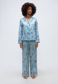 Γυναίκα που στέκεται ξυπόλητη φορώντας ανοιχτό μπλε ριγέ πιτζάμες με πολύχρωμα τυπώματα αρκουδάκια και μακριά μανίκια και παντελόνια.