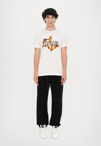 Balta marškinėliai su juodais ir oranžiniais "Versace Jeans Couture" logotipais. Juodos kelnės su tekstūruotu logotipo raštu. Baltos sportbačiai su juodais akcentais.
