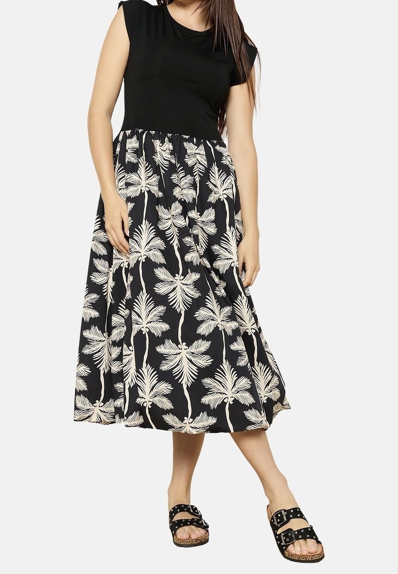 Femme portant une robe noire sans manches avec un motif de palmier blanc sur la jupe, associée à des sandales noires à boucle.