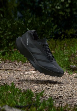 Chaussure de trail running Adidas Terrex noire en l'air au-dessus d'un chemin rocheux avec un fond de feuillage vert.