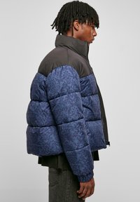 Prešívaná puffer bunda v tmavomodrej farbe s tonálnym kvetinovým vzorom a čiernou vrchnou časťou. Obsahuje vysoký golier a zips. Štruktúrovaná látka.