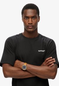 Zwarte katoenen t-shirt met het "SUPERDRY" logo in het wit op de borst, gecombineerd met een goudkleurig horloge met een bruine leren band.