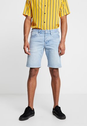Homme portant un short en denim bleu clair, une chemise jaune à manches courtes avec des rayures noires et des baskets noires, debout devant un fond uni.