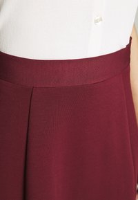 Burgundivärgad kjol med en slät textur, försedd med en bred midjeband och veckdetaljer, tillsammans med en vit knapp-up topp.