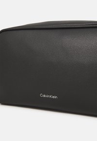 Svart kosmetikkveske i lær med teksturert overflate og sølvfarget "Calvin Klein"-logo. Rektangulær form med glatte kanter og minimalt design.