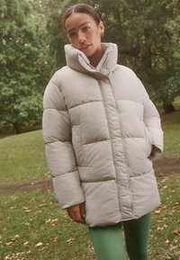 Lys beige polstret jakke med høj krave og forreste trykknapper, der har en boxy form og synlig quiltedesign.