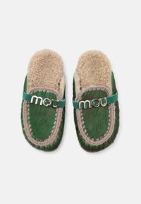 Green fur slippers with a beige sheep wool lining, featuring decorative stitching and a green band with a gold "mou" logo.