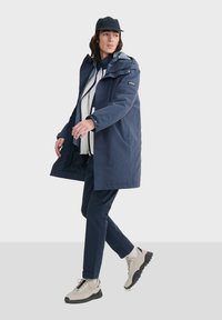 Manteau imperméable bleu marine avec fermeture zippée, capuche et doublure intérieure à motifs, associé à un pantalon bleu marine et des chaussures claires.