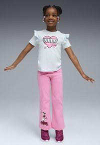 Niña pequeña de pie con los brazos extendidos, vistiendo una camiseta de manga rizada azul claro de Puma y pantalones rosas con personajes de dibujos animados, además de zapatillas rosas.