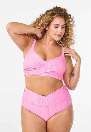 Frau mit lockigem Haar, die den Riemen eines gerippten rosa Bikinis mit Wickeldesign anpasst, stehend vor einem weißen Hintergrund.