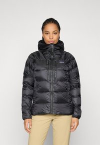 Vrouw met vlechten die een zwarte pufferjack met capuchon en beige broek draagt, staande tegen een effen lichte achtergrond.
