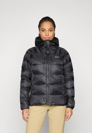 Vrouw met vlechten die een zwarte pufferjack met capuchon en beige broek draagt, staande tegen een effen lichte achtergrond.