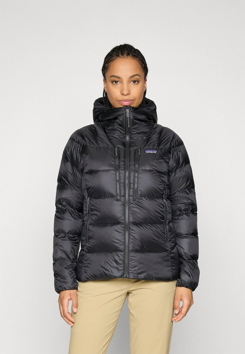 Vrouw met vlechten die een zwarte pufferjack met capuchon en beige broek draagt, staande tegen een effen lichte achtergrond.
