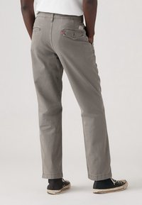 Grå bomullstwillbyxor med rak ben, två bakfickor och ett synligt rött logotag. Bärs med svart-vita sneakers.