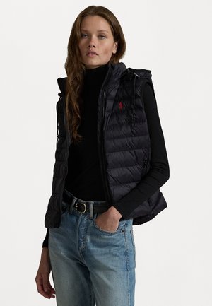 Jonge vrouw in een zwart gewatteerd mouwloos jack over een zwart coltrui en lichtblauwe jeans, staande met één hand in haar zak.