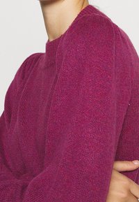 Pull en tricot fuchsia avec un col rond et des poignets côtelés. Texture douce et léger éclat, présentant des variations subtiles de couleur.