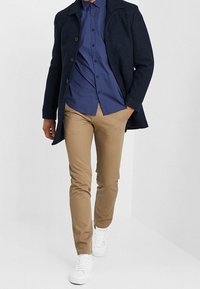 Marinblå ylleblandad kappa, mönstrad blå skjorta, beigefärgade slim fit-byxor och vita sneakers. Kappan har krage med vecko och knappar.