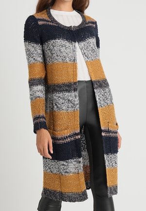 Vrouw draagt een lange, meerkleurige gebreide cardigan met horizontale strepen in mosterdgeel, marineblauw, grijs en wit, gecombineerd met een zwarte broek en een wit shirt.