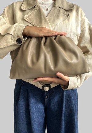 Borsa a tracolla in pelle beige con design arricciato, tenuta da una persona che indossa una giacca chiara e jeans blu scuro. Texture liscia.